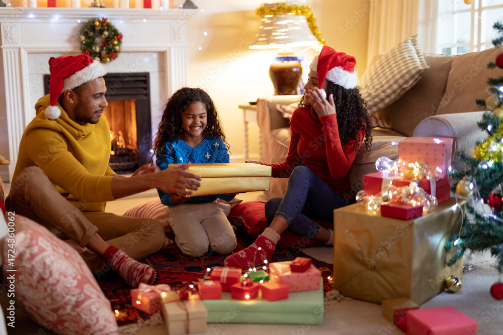 Fototapeta premium African American family in Santa hats exchanging gifts on rug in living room near Christmas tree