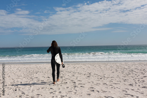 Female surfer in black wetsuit walking on beach carrying white surfboard and leash toward waves
