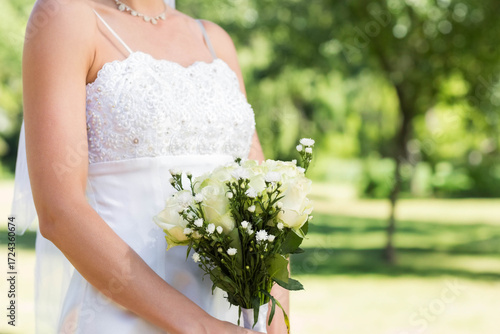 White wedding gown resting on garden lawn, holding white rose bouquet accented by greenery