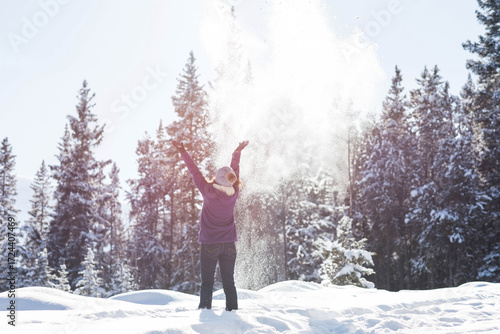 Mid adult woman wearing purple jacket, standing in deep snowdrift in pine forest, tossing snow