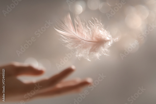 A soft, delicate feather floats gently above an outstretched hand against a blurred, neutral background with subtle light reflections