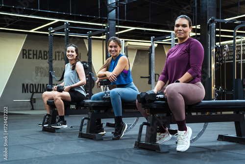 Three latin american women smiling, sitting on benches in a gym, holding dumbbells, showing fitness and wellness