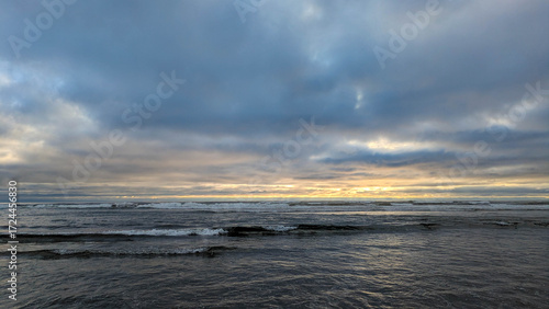 Side view from a wet beach with an emphasis on the small waves rolling in, out westward towards the pacific ocean, under a majestic and complicated cloud-scape near dusk. 