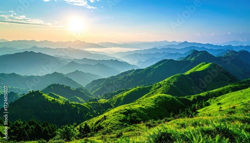 Lush, grassy mountaintop overlooking rolling, forested hills under a bright blue sky with the sun rising in the distance