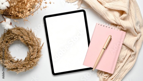 White backdrop with a tablet (blank screen), pink notebook with gold pen, wreath, bowl of dried plants, and a soft, textured scarf