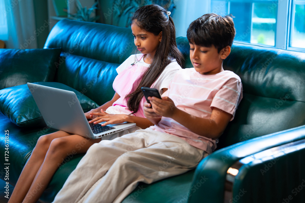 Obraz premium Indian kids sitting on sofa using laptop and smartphone, siblings enjoy digital time at home