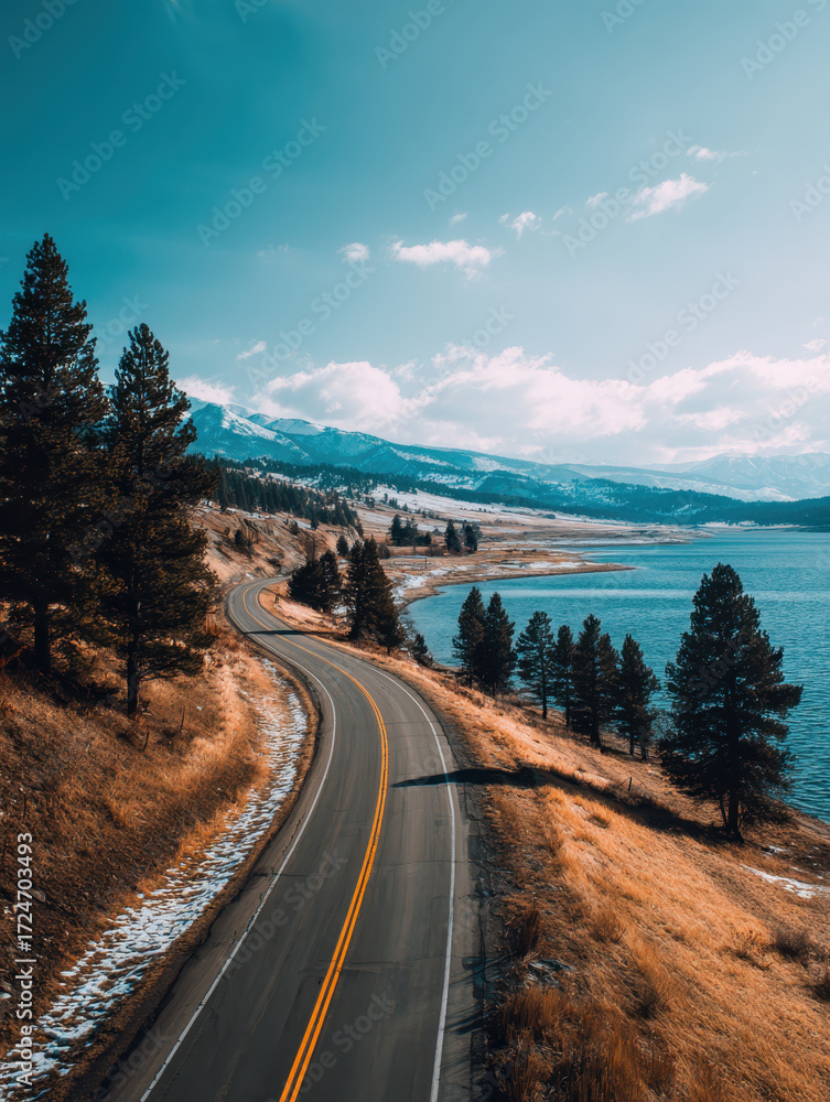 Fototapeta premium Winding road curves along serene lakeside, bordered by tall pine trees and golden grass under clear blue sky. Snow capped mountains rise in distance, creating tranquil landscape