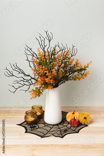 A white vase with autumn branches and Halloween decor stands on a wooden table. Mini pumpkins, witch hats, a broom, and a spiderweb cloth create a festive seasonal atmosphere.