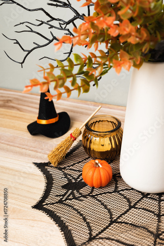 A white vase with autumn branches and Halloween decor stands on a wooden table. Mini pumpkins, witch hats, a broom, and a spiderweb cloth create a festive seasonal atmosphere.