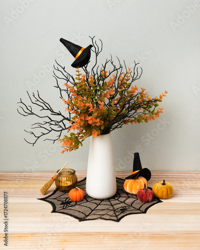 A white vase with autumn branches and Halloween decor stands on a wooden table. Mini pumpkins, witch hats, a broom, and a spiderweb cloth create a festive seasonal atmosphere.