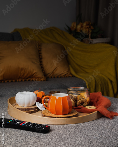 A cozy autumn scene with a tray on a sofa featuring a pumpkin-shaped mug of coffee, cookies in a glass jar, and decorative candles. Warm pillows and a blanket create a relaxed seasonal atmosphere.