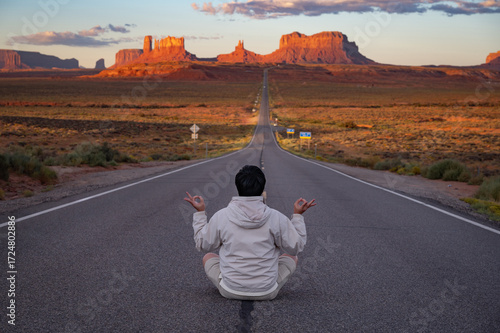 Rear view of Asian man tourist sitting on highway US route 163 looking at the long road view leading to Monument Valley on the Arizona-Utah border at sunrise , United States. America road trip travel
