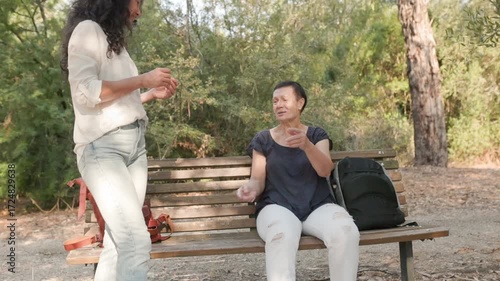 Senior woman in navy blouse and white pants gesturing expressively while sitting on park bench with black backpack, younger woman in white blouse standing nearby, communication and connection.