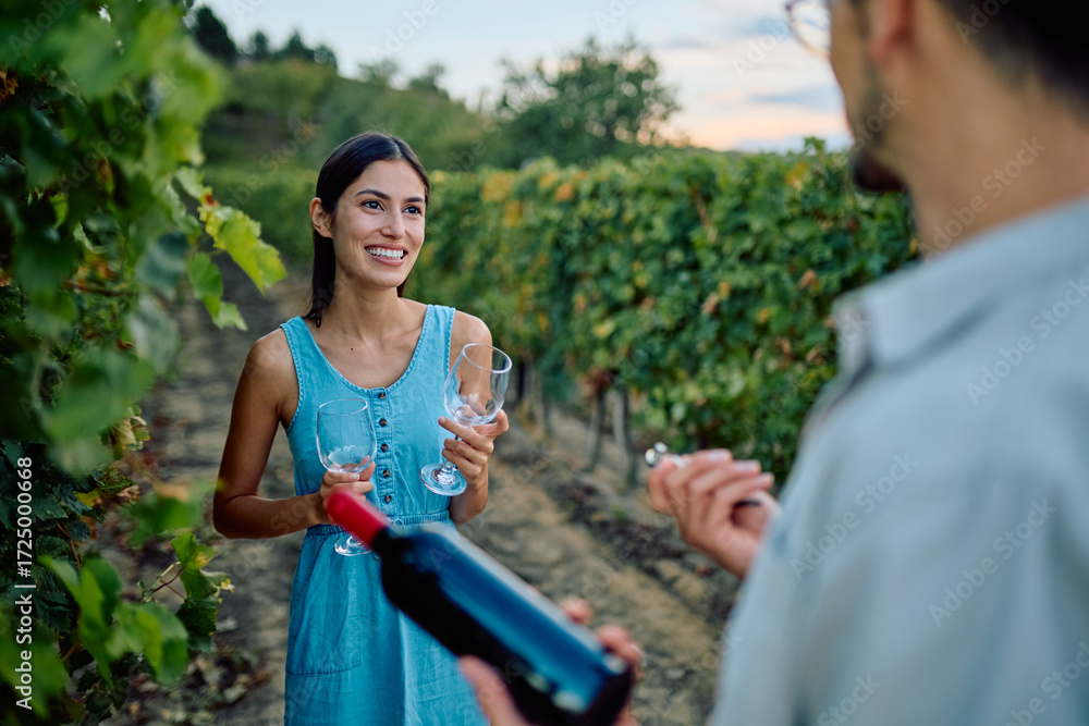 Obraz premium Young woman smiling, holding wine glasses, while man prepares to open a bottle of red wine in a scenic vineyard