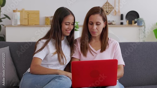 Young teenage student girl sitting on sofa at home with her mid adult mother, both sharing time together while using laptop computer. Family and technology lifestyle concept