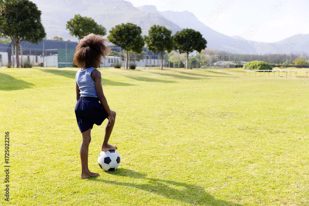 Obraz premium African American girl in sportswear standing barefoot on sports field resting foot on soccer ball