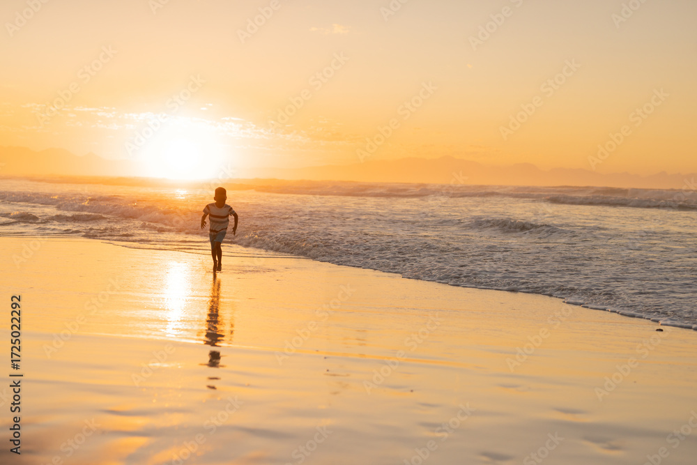 Fototapeta premium African American boy running at sunset on wet beach sand, waves lapping and sun reflecting