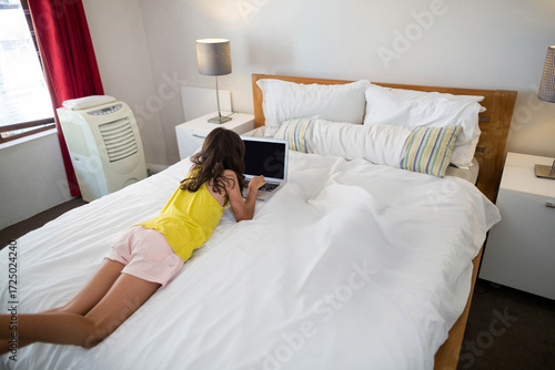 Child lying on stomach across wooden bed using laptop in bedroom with red curtains and lamps