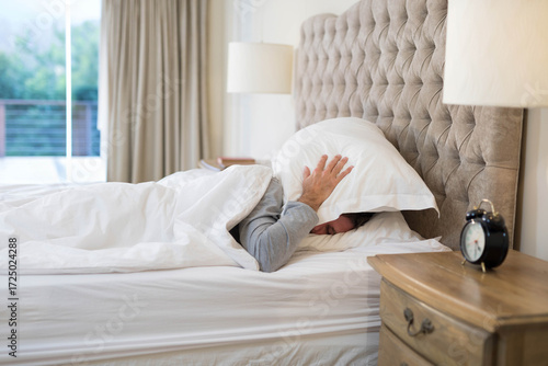 Man lying under white duvet in bedroom covering head with pillow on bed beside nightstand lamp