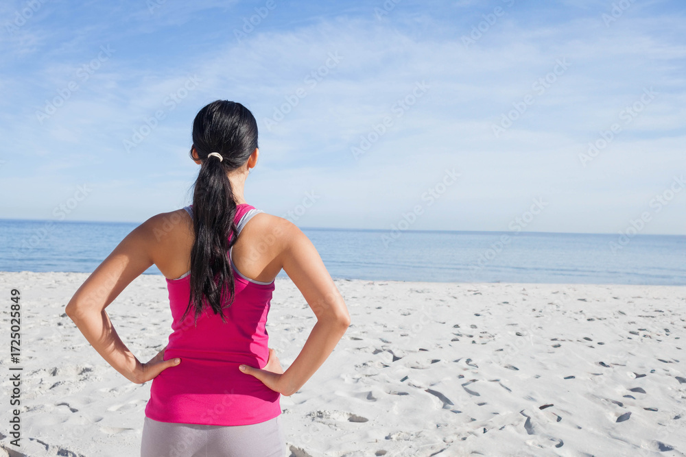 Naklejka premium Asian woman standing on white sand beach facing calm blue sea wearing pink top and leggings