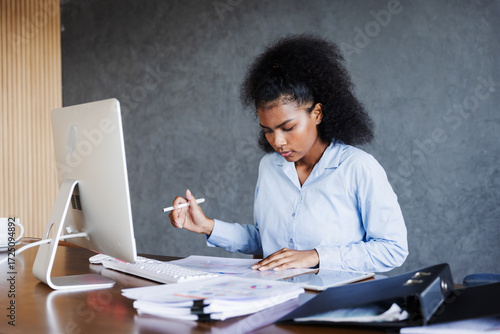 A young professional woman works at her desk with a computer, reviewing charts and opening a file folder, reflecting focus, organization, productivity, and dedication in a modern business environment.