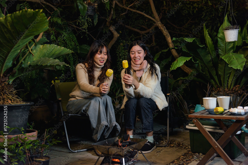 Obraz premium Happy women friends enjoy winter outdoor camp forest night, people gather warm fire roasting corn in cozy sweaters, creating cheerful winter memories