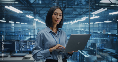 Portrait of a Data Center Administrator Standing in a High-Tech Server Room, Optimizing Server Configurations on a Laptop Computer. Female Specialists Looking at Camera and Smiles