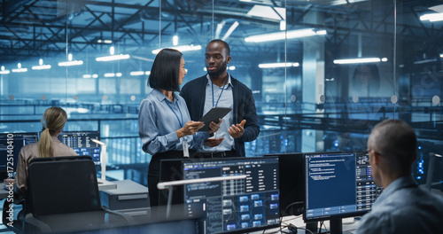 Male DevOps Engineer and Female IT Consultant Using a Tablet Computer, Standing in an Office Above the Rows of Servers in a Data Center, Developing Automation Scripts for Artificial Intelligence