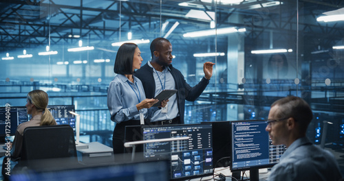 Male DevOps Engineer and Female IT Consultant Using a Tablet Computer, Standing in an Office Above the Rows of Servers in a Data Center, Developing Automation Scripts for Artificial Intelligence