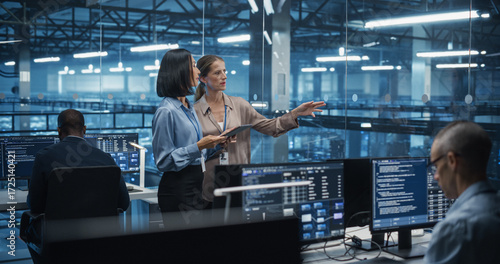 Two Diverse Female Cloud Security Engineers Work Side by Side on a Tablet Computer in a Data Center Office, Conducting a Thorough Security Audit of the Server Network Infrastructure