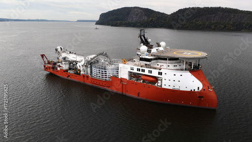Nyack, United States - 16 September 2025: Aerial view of a vibrant red and white offshore support vessel sailing on the Hudson River, framed by the lush, green shores of the Palisades cliffs.