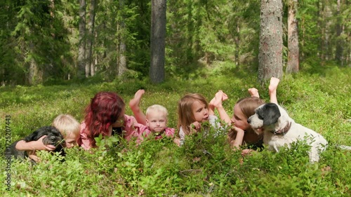 Five children and two dogs lie on a sunlit forest clearing in slow motion, laughing, waving, and enjoying nature on a carefree summer day