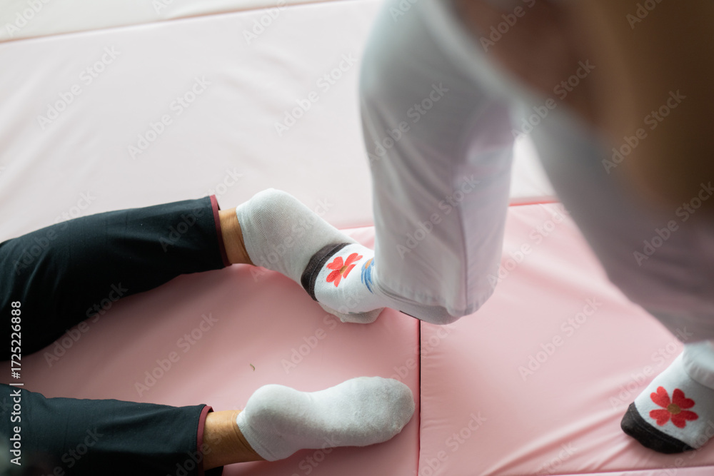 Obraz premium Close up of physiotherapist assisting an older woman by holding her legs during a rehabilitation exercise on a mat. Physiotherapy session concept emphasizing mobility recovery, strength training, and