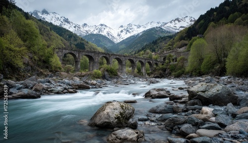 A serene mountain river flows beneath a historic stone arch bridge, framed by lush green vegetation and snow-capped peaks.