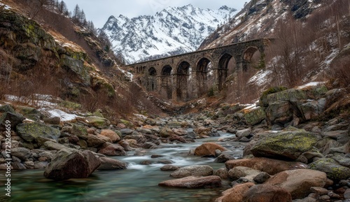 A serene mountain river flows past an ancient stone arch bridge, nestled amongst snowy peaks.