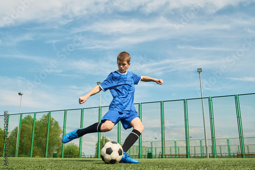 Teenage boy kicking soccer ball on outdoor field, wearing sports uniform, showing athletic movement with focused expression, green fence and blue sky in background