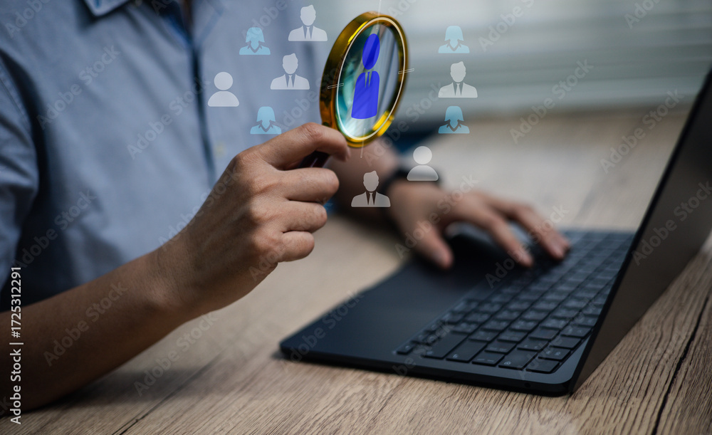 Fototapeta premium Hand holding magnifying glass in front of laptop with digital human icons. Concept of HR recruitment, talent search, employee hiring, business workforce, and candidate selection