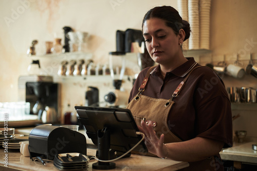 Caucasian young adult woman working behind counter using touchscreen point of sale terminal in coffee shop, standing near stacked cups and kitchen equipment, focused on transaction