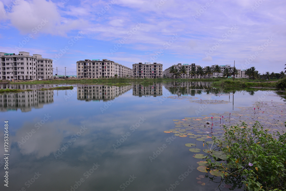 Fototapeta premium Modern residential apartment buildings reflected in a calm pond, with blooming wildflowers and greenery in the foreground. A scenic blend of urban development and natural beauty in Bangladesh
