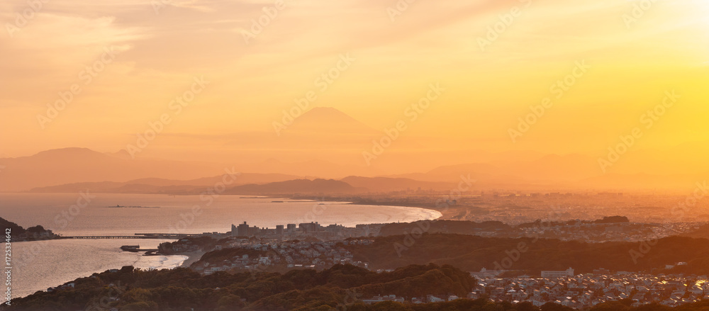 Aerial view of the coastline meeting lush hills, the city sprawling towards the horizon, and Mount Fuji veiled in the warm glow of the setting sun, Kamakura, Kanagawa, Japan.