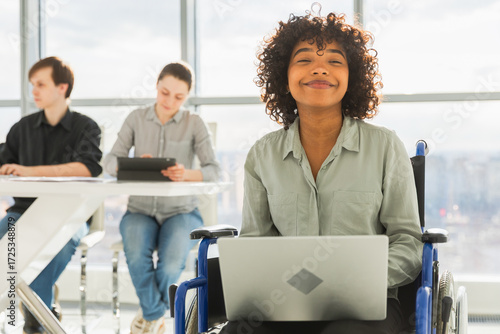 Accessible environment. African woman in chair for people with disability working with laptop in modern office. Diverse multiracial group of people colleagues working together. Young professional team