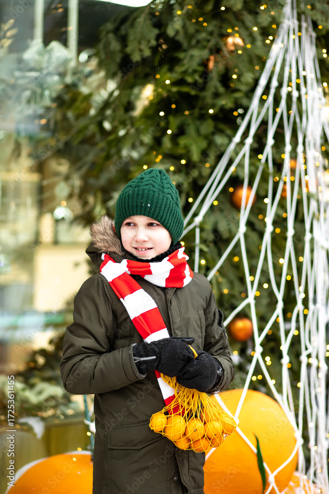 Obraz premium Happy cute teenager boy with mesh bag of tangerines standing near by spruce Christmas tree. Holiday celebrate concept.