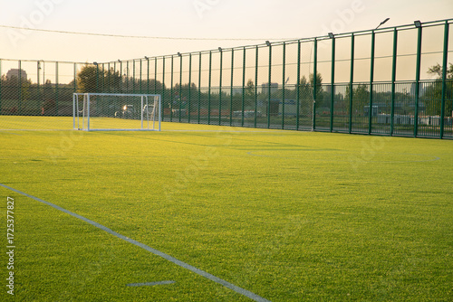 Green artificial soccer field in late afternoon with goalposts and fencing. English football pitch