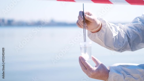 Person in protective hazmat suit and medical mask conducting water testing near lake. Environmental pollution research, contamination control, biohazard investigation, and safety inspection outdoors.
