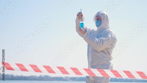 Person in protective hazmat suit and medical mask conducting water testing near lake. Environmental pollution research, contamination control, biohazard investigation, and safety inspection outdoors.