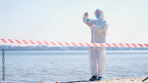 Person in protective hazmat suit and medical mask conducting water testing near lake. Environmental pollution research, contamination control, biohazard investigation, and safety inspection outdoors.