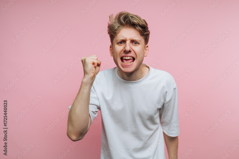 Fototapeta premium A young man in a white t-shirt expressing anger with a fist raised against a pink background.