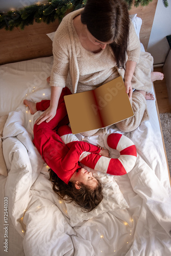 Mother and child are relaxing in bed on Christmas morning, enjoying quality family time while reading a festive storybook, creating a cozy and loving holiday atmosphere