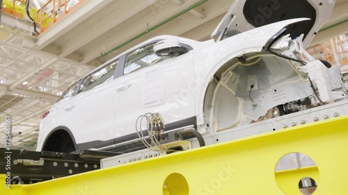 The undercarriage of a car body moves along an assembly line conveyor in an automobile factory. A low angle view from beneath the industrial conveyor belt with a protective metal grate.