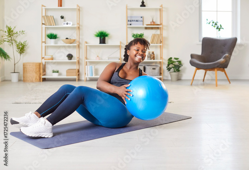 Schilderij op canvas Positive smiling young African American woman doing intense workout on yoga mat in living room at home, performing russian twist exercise with fitball for weight loss, core and abdominal muscles